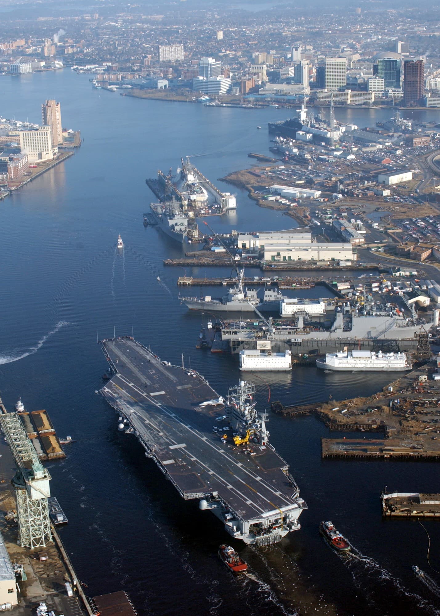An aerial view of the Elizabeth River at Norfolk Naval Shipyard, with downtown Norfolk in the background and an aircraft carrier transiting the river